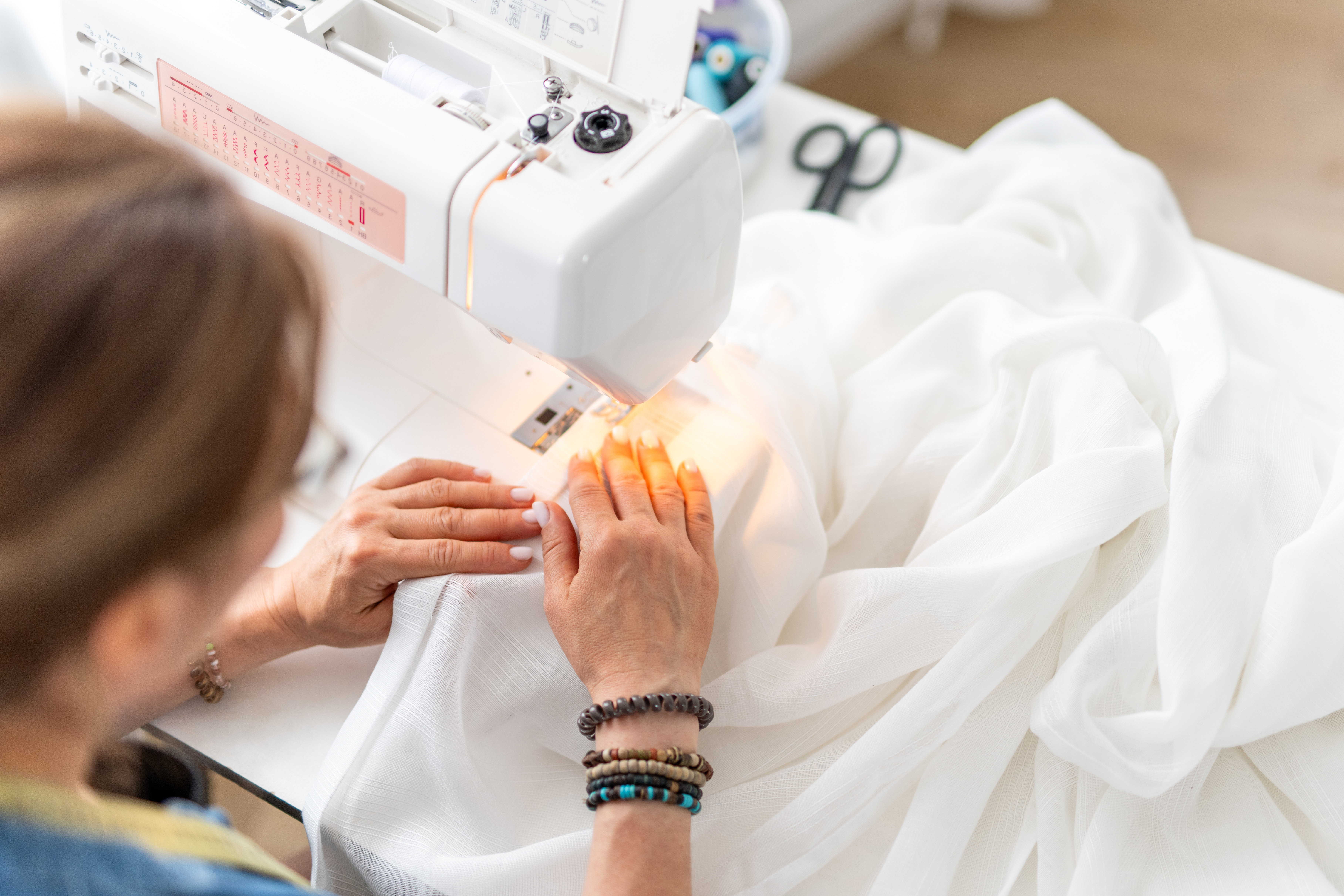 Woman operates sewing machine, stitching white fabric on tabletop. Hands guide material.